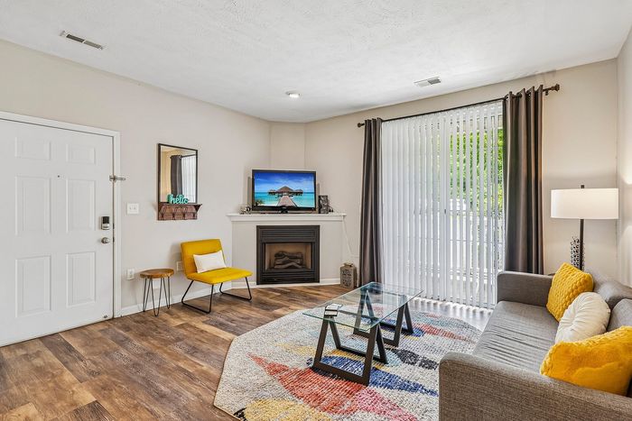 Inviting living room with modern decor, featuring a cozy gray sofa, vibrant yellow accents, and a sleek glass coffee table on a colorful rug, illuminated by natural light streaming through large vertical blinds.