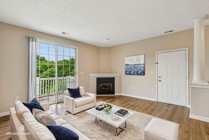 Bright, virtually staged living room with cozy fireplace, elegant column, and large sliding glass doors opening to lush greenery.