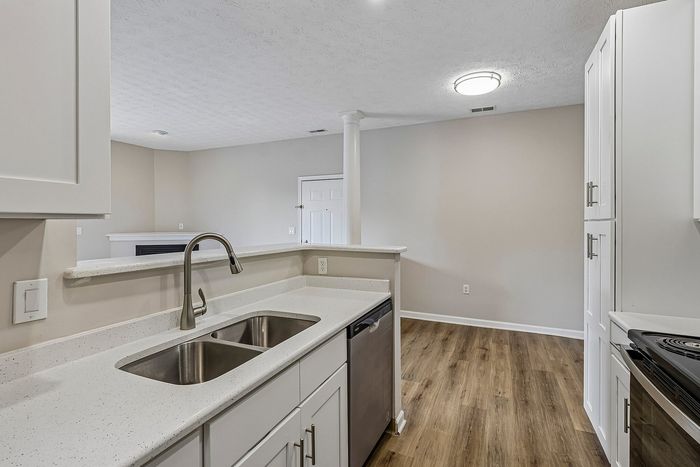 Modern kitchen with sleek white cabinetry, elegant countertops, and stainless steel appliances, overlooking a bright living area with wooden flooring.