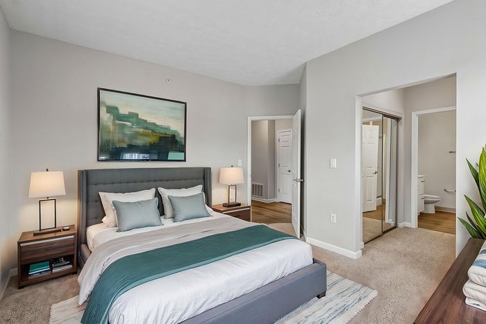 Elegant bedroom with modern decor, featuring a plush gray bed, soft lighting, and a stylish painting above; mirrored closet doors add spaciousness, blending comfort and sophistication.