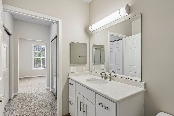 Elegant bathroom with a modern vanity, bright lighting, and a spacious mirror, leading into a cozy carpeted bedroom with a large window.