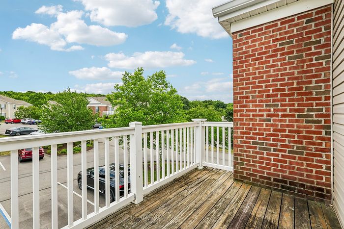 Charming balcony with a wooden deck and white railing, overlooking a scenic residential view with lush trees and clear skies.