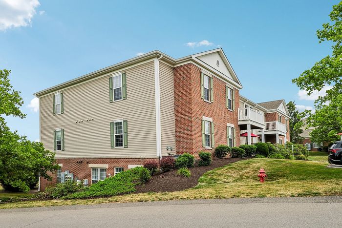 Charming brick apartment building with green shutters nestled in lush landscaping, featuring balconies and a peaceful neighborhood setting under a clear blue sky.