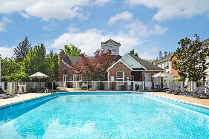 Inviting outdoor pool area with clear blue water, surrounded by a charming brick clubhouse and lush greenery, perfect for relaxation.