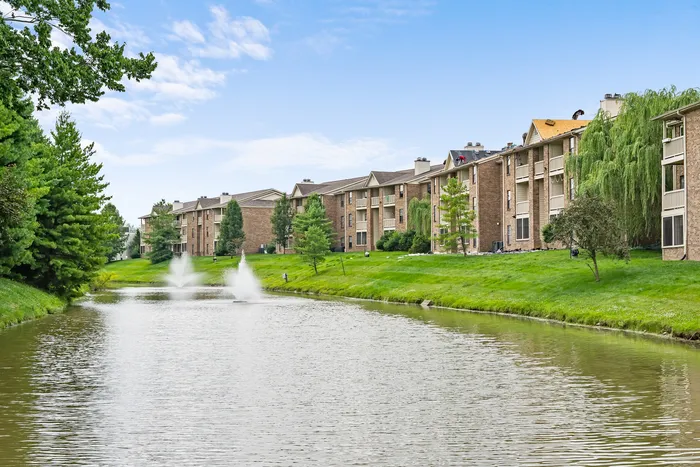 Charming apartment complex nestled by a serene pond with fountains and lush greenery.
