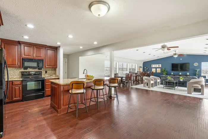 Elegant open-concept living area featuring a spacious kitchen with cherry wood cabinets, a stylish granite island with bar seating, and a cozy living room with modern furnishings and vibrant accent wall.