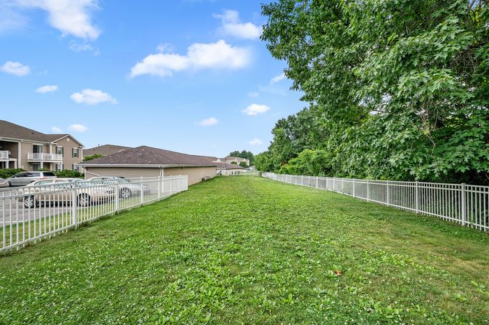 Expansive green backyard with white fencing, adjacent to charming residential buildings and lush trees, offering a serene and secure environment.