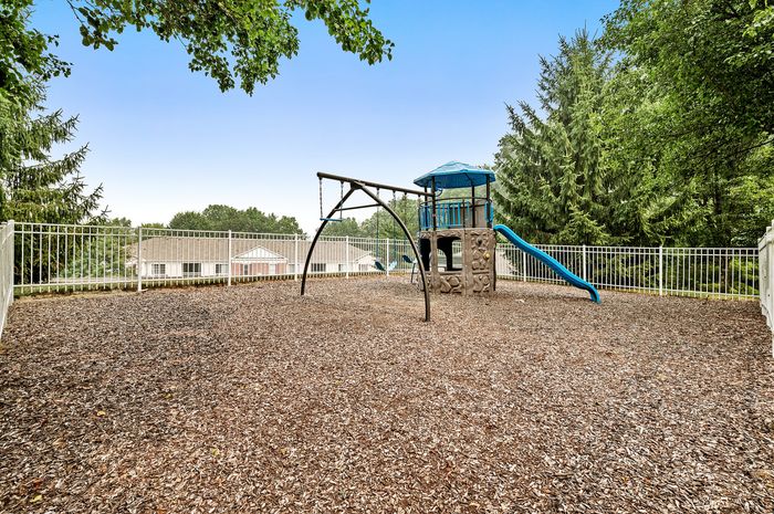 Inviting playground surrounded by lush greenery, featuring swings, a climbing structure, and a vibrant blue slide within a safe gated area.