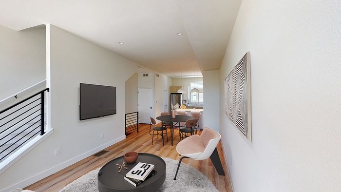 Open-concept living room adjacent to a dining area with a modern dining table and kitchen in the background, featuring wood-style flooring and large windows