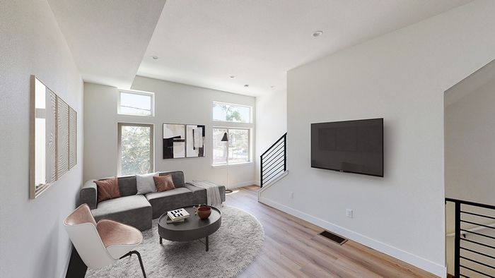 Living room with sleek gray couch, fluffy rug, wood-style flooring, wall-mounted TV, large bright windows, and high ceilings