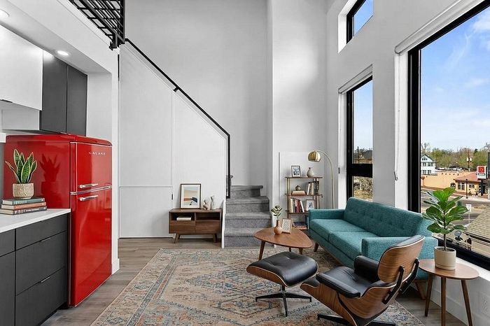 Chic and modern living room with a vibrant red retro fridge, stylish teal sofa, elegant staircase, and expansive windows offering a scenic view.