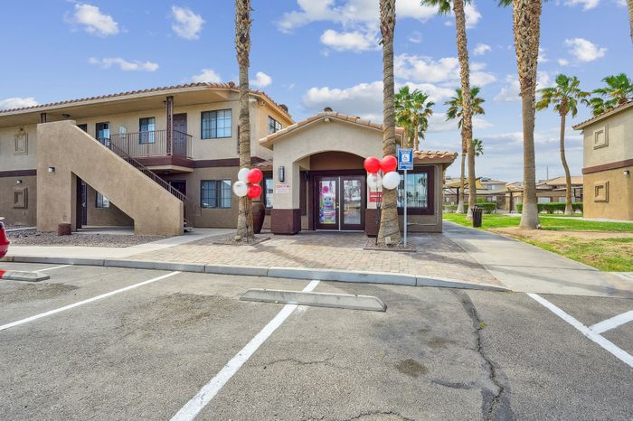 Inviting apartment complex entrance with festive red and white balloons, palm tree landscaping, and sunny skies.