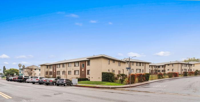 Charming corner apartment complex with well-maintained landscaping and ample parking, set against a clear blue sky.