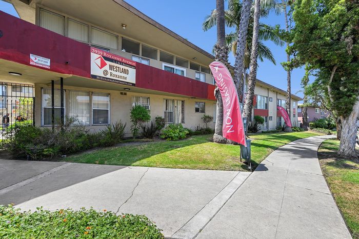 Inviting apartment complex with lush greenery and vibrant 'Now Leasing' signs, offering comfortable living on Roxanne Avenue.