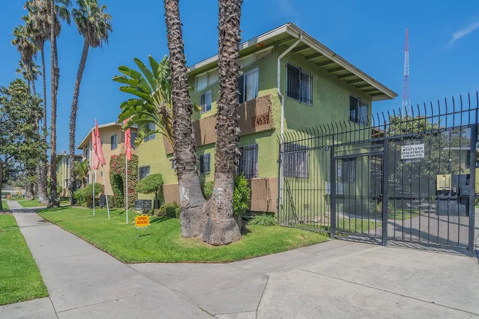 Charming suburban apartment building with lush landscaping and vibrant greenery, highlighted by tall palm trees and a secure gated entrance.