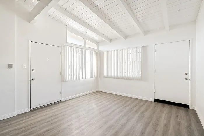 Bright and airy room with vaulted ceiling, featuring modern wood flooring and large windows with vertical blinds.