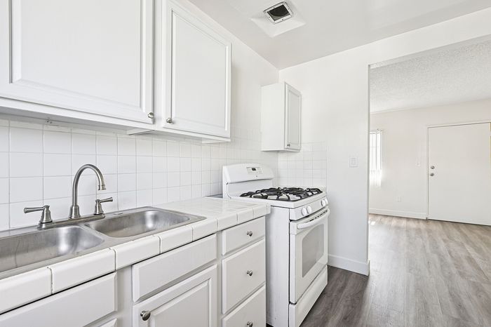 Bright kitchen with modern white cabinetry, dual stainless steel sink, and an efficient gas stove, leading into a sunlit open living space with elegant flooring.