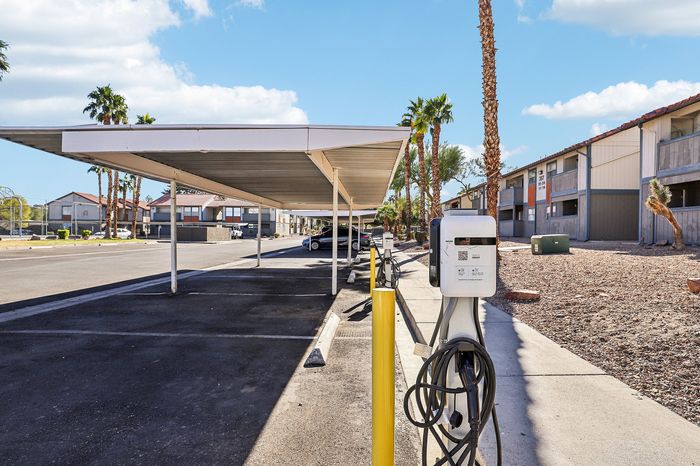 Modern apartment complex with covered parking and electric vehicle charging station under sunny skies, surrounded by palms.