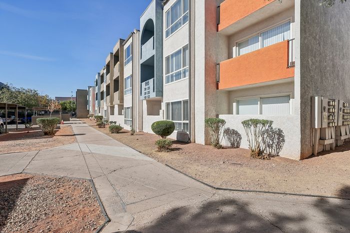Vibrant, modern apartment complex exterior with colorful facades, large windows, and lush greenery along a welcoming, sunlit pathway.