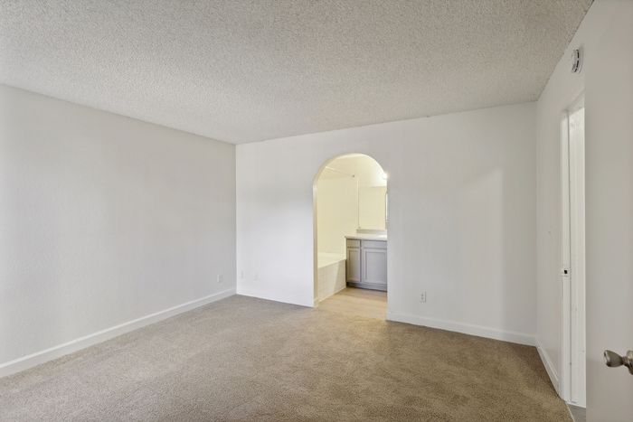 Bright and spacious room with plush carpeting and an elegant archway leading to a modern bathroom, offering comfort and style.