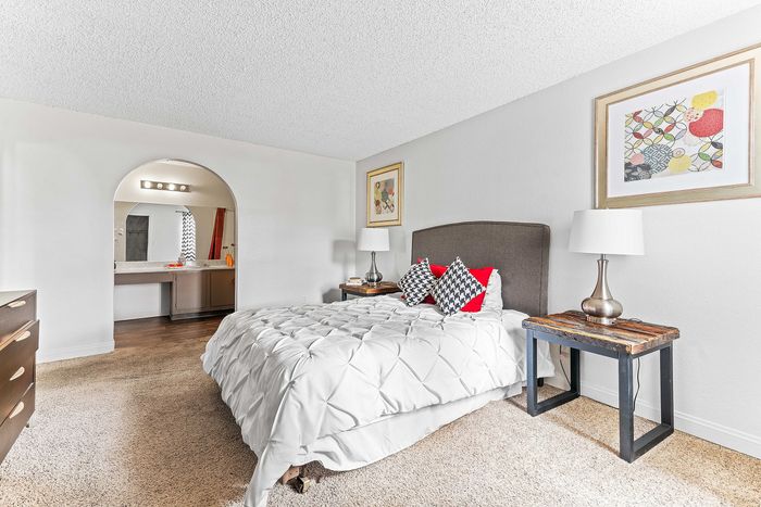 Bright and inviting bedroom featuring a plush bed with checkered and red accent pillows, a rustic bedside table with a modern lamp, and an elegant vanity space in the background.