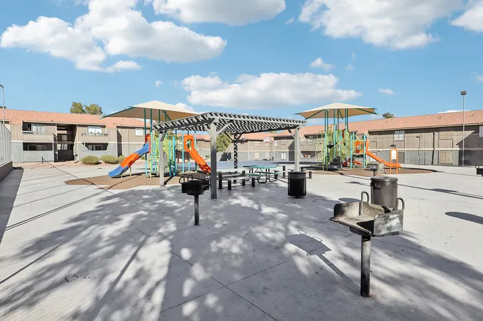 Charming apartment outdoor area featuring colorful playground with slides, shaded picnic tables, and barbecue grills under a vibrant blue sky.