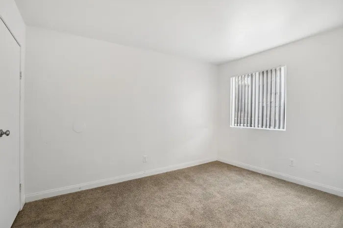 Bright and airy bedroom featuring soft carpet flooring and a large window with vertical blinds, perfect for cozy living.