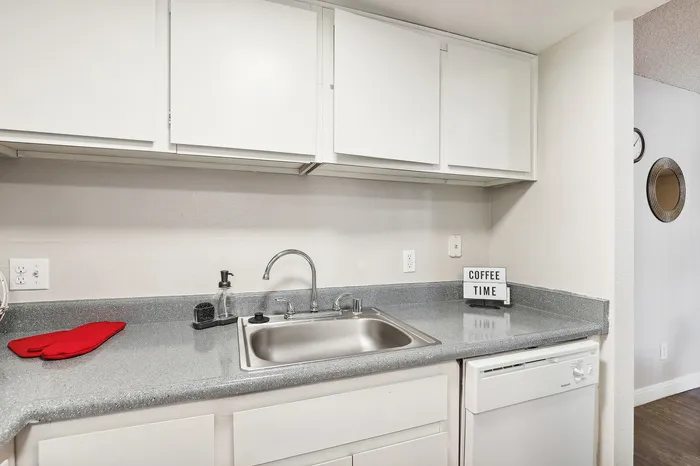 Contemporary kitchen with sleek gray countertops, stainless steel sink, and white cabinetry, featuring a modern dishwasher and charming 'Coffee Time' sign for a cozy atmosphere.