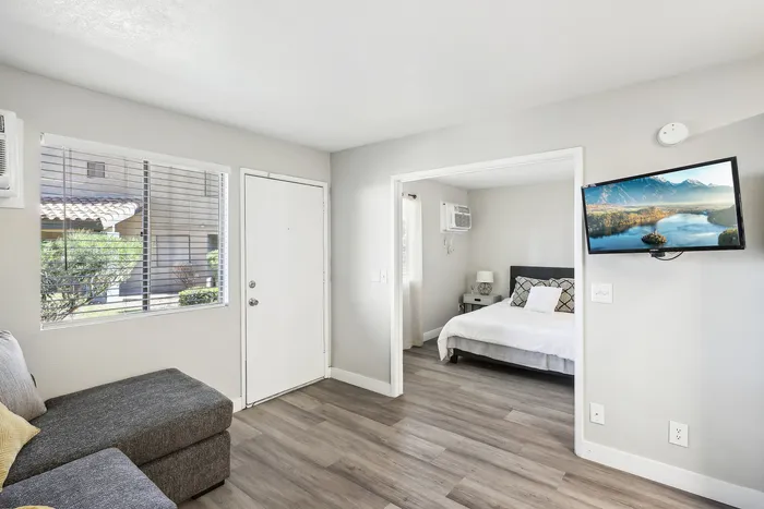 Bright and modern living area with a sectional sofa, wall-mounted TV, and a glimpse into a cozy bedroom; large windows offer a view of the serene outdoor space.