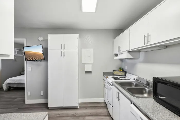Modern kitchen with sleek white cabinetry, dual sink, and built-in stove; cozy bedroom glimpse with natural light and wood flooring.