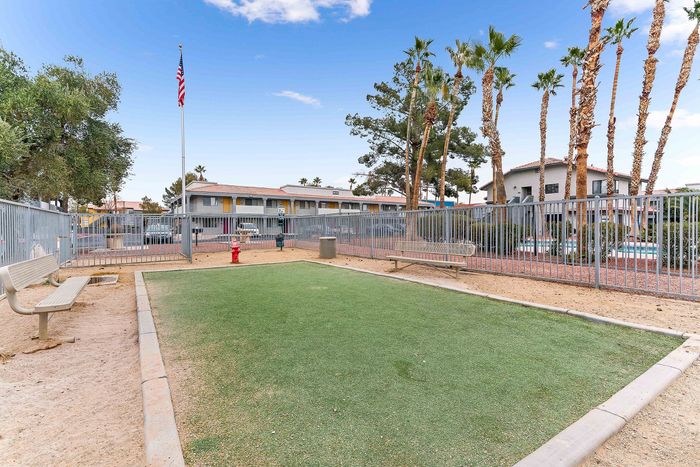 Inviting apartment complex with a serene outdoor area featuring a manicured green space, benches, and vibrant palm trees against a clear blue sky.