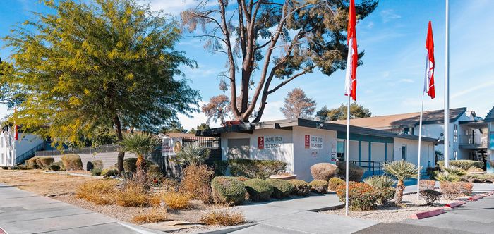 Charming apartment community with lush landscaping, vibrant flags, and modern leasing office under a clear blue sky.