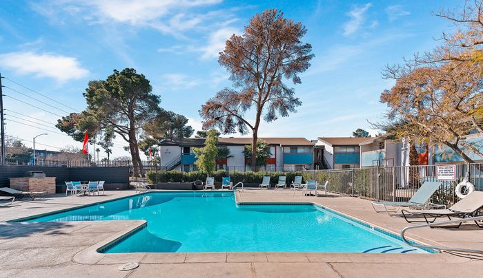 Inviting apartment complex with a sparkling swimming pool, spacious deck, and lush trees under a clear blue sky.