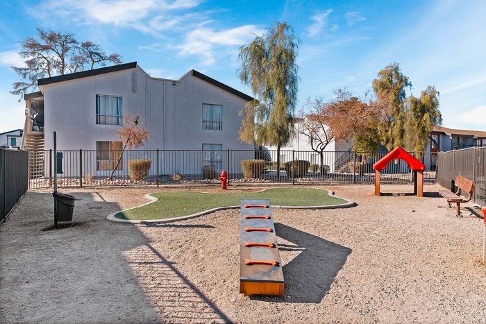 Charming apartment complex with a private playground featuring green landscaping, a bench, and climbing equipment under a bright blue sky.