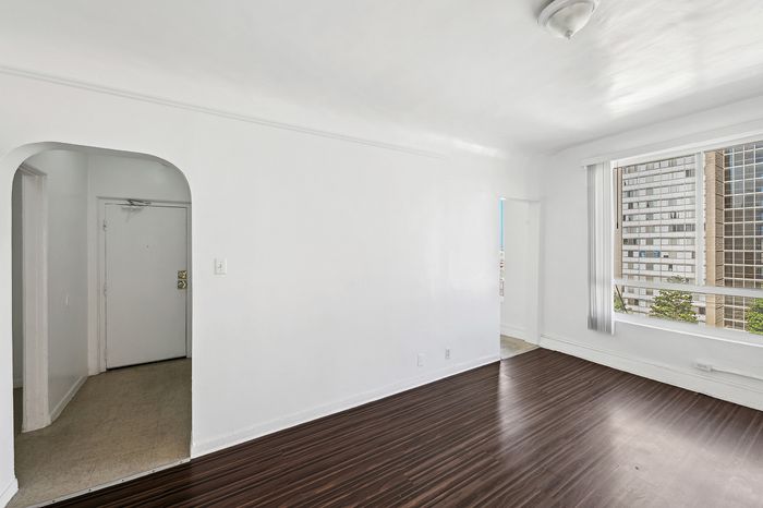 Bright open living space with sleek hardwood floors and a large window offering city views in a modern apartment.