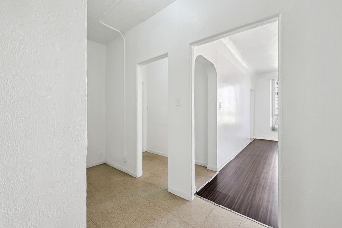 Bright and inviting hallway featuring crisp white walls, leading to a spacious room with elegant wood flooring, offering modern living charm.