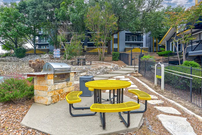 Charming outdoor area with vibrant yellow picnic table, BBQ grill, and lush surroundings, perfect for gatherings.