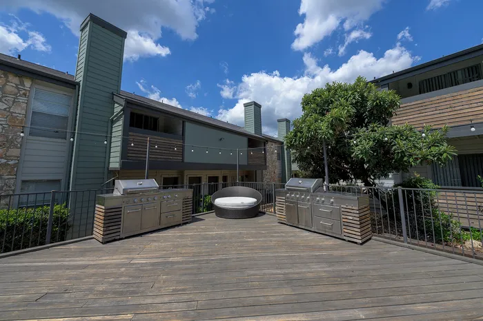 Chic apartment exterior with a spacious wooden deck, featuring modern stainless steel grills, a cozy round seating area, lush greenery, and string lights under a vibrant blue sky.