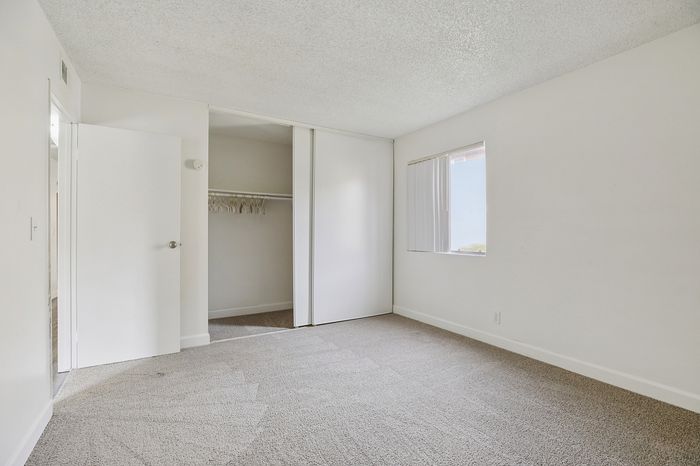 Bright and spacious bedroom with fresh carpeting, large closet, and natural light from a window with vertical blinds.