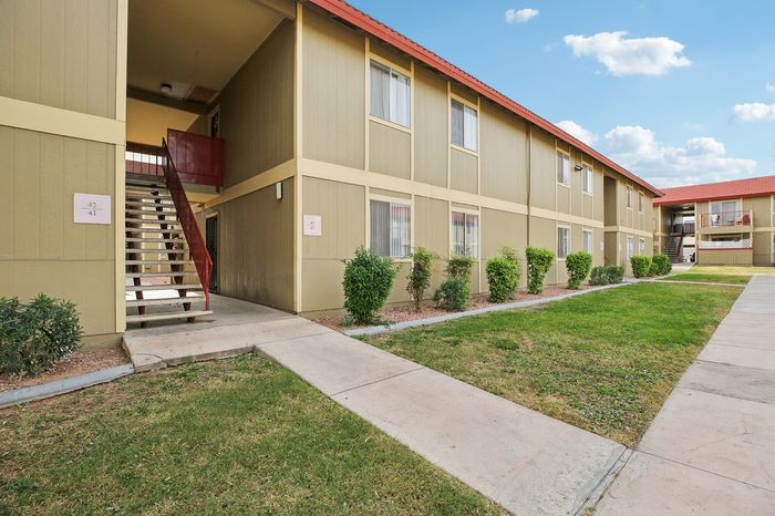 Charming apartment complex with a vibrant red roof, surrounded by lush greenery and a well-maintained walkway under a clear blue sky.