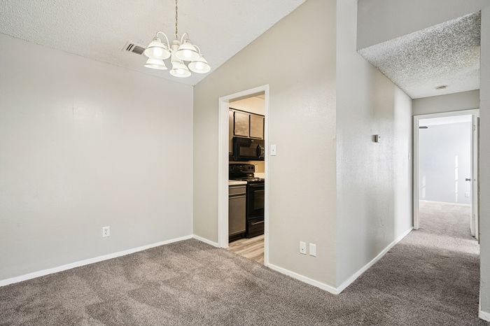 Bright apartment interior featuring a cozy carpeted dining area with elegant overhead lighting, adjacent to a modern kitchen and a sunlit hallway leading to bedrooms.