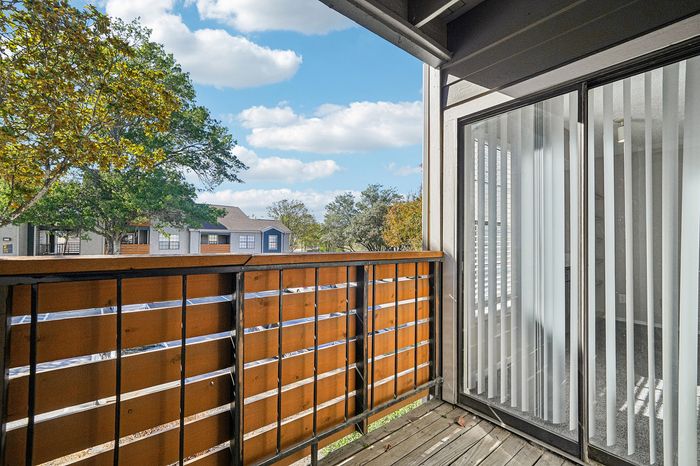 Charming balcony with wooden railing offering a scenic view of lush trees and residential buildings under a bright blue sky.