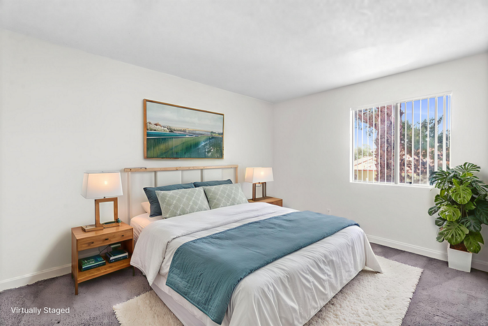Bright and inviting bedroom featuring a cozy bed with elegant blue and white bedding, large window with natural light, modern lamps on wooden nightstands, and a potted plant for fresh ambiance.