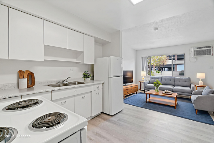Bright and modern open-plan living area featuring a sleek kitchen with white cabinetry, cozy living space with stylish gray sofa, large window for natural light, and elegant decor. Perfect for comfortable living.
