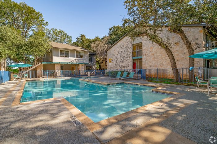 Inviting apartment complex with a sparkling pool, surrounded by lush trees and charming stone buildings, offering a serene outdoor retreat.