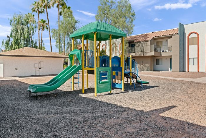 Vibrant playground area with colorful slides and climbing equipment beside modern apartments, complemented by tall palm trees and a clear blue sky.