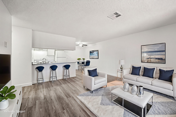 Contemporary open-plan living room with stylish white furnishings, elegant accent pillows, and a sleek kitchen with a breakfast bar and modern stools, creating a welcoming and chic atmosphere.
