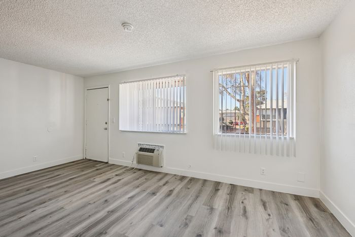 Bright and airy living space with modern light wood flooring, two large windows allowing natural light, and a sleek air conditioning unit.