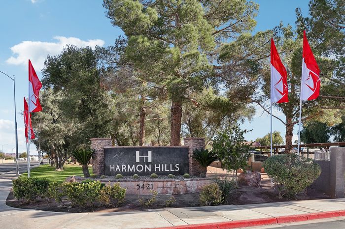 Inviting entrance to Harmon Hills with vibrant red and white flags surrounded by lush greenery and tall trees, offering a serene living environment.