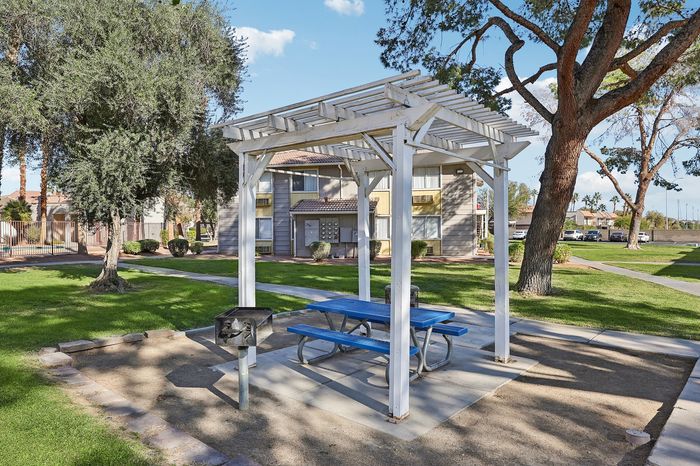Inviting community picnic area with a pergola, blue picnic table, and barbecue grill, set amidst lush green gardens and charming residential buildings.